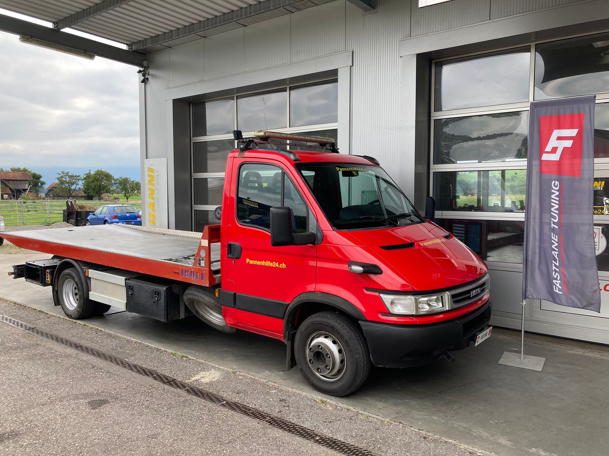 Ein leuchtend roter IVECO Dailiy Abschleppwagen parkt vor der Garage neben der Fastlane Tuning Schweiz Flagge.