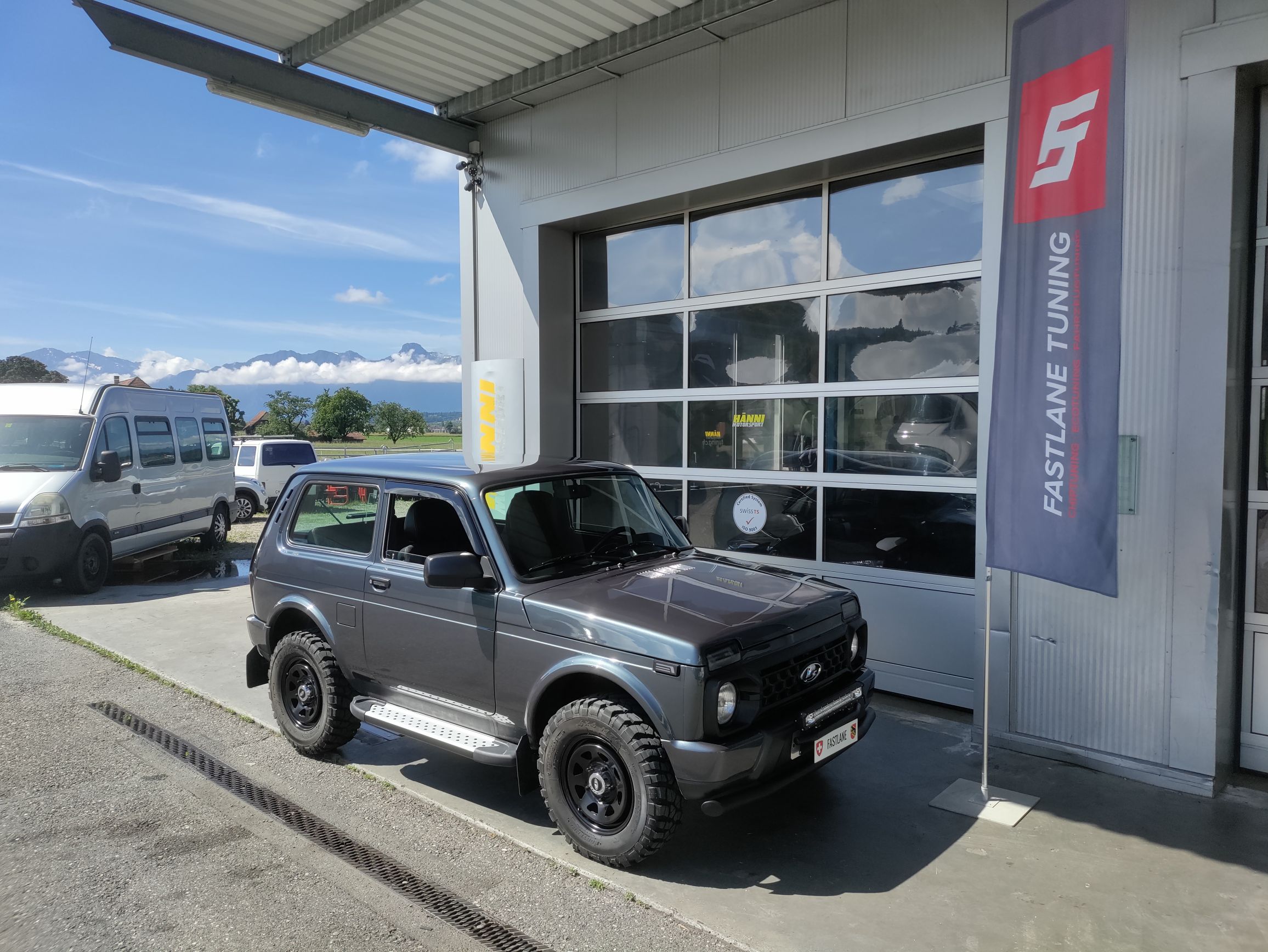 Ein kleiner grauer Lada 4 X 4 parkt vor der Garage neben der Fastlane Tuning Schweiz Flagge.