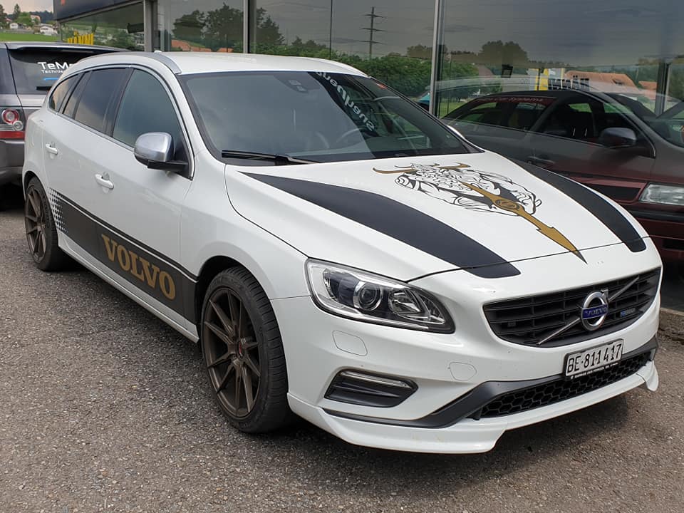 Ein weiß-schwarzer Volvo T6 Kombi mit einem Motiv des nordischen Gottes auf der Motorhaube. Er ist neben einem Autohaus geparkt.