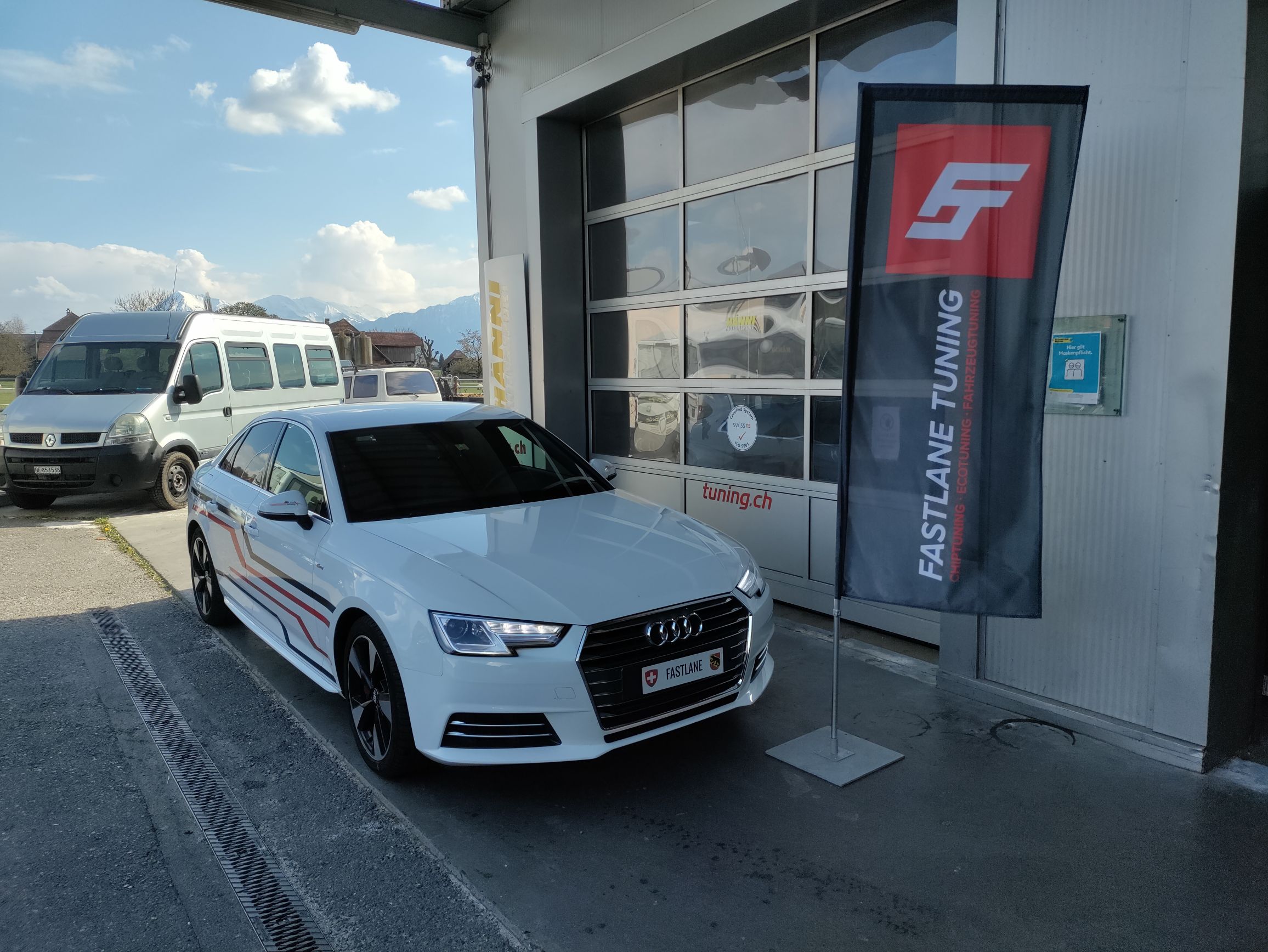 Ein weisser Audi A4 35 TFSI mit roten und schwarzen Streifen an den Seiten, geparkt vor der Garage neben der Fastlane Tuning Schweiz Flagge