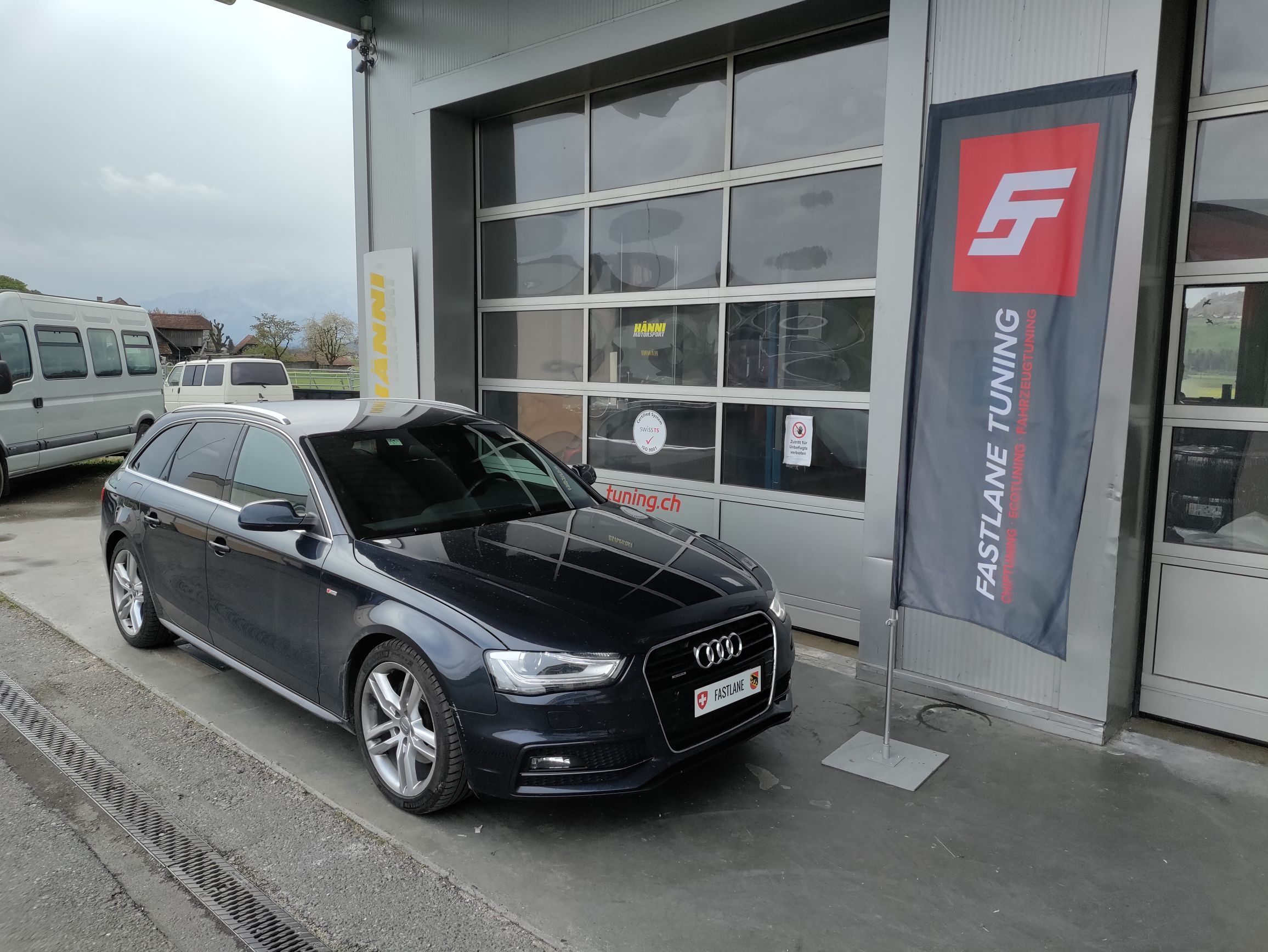 Ein schwarzer Audi A4 Kombi 2.0 TDI vor der Garage neben der Fastlane Tuning Schweiz Flagge