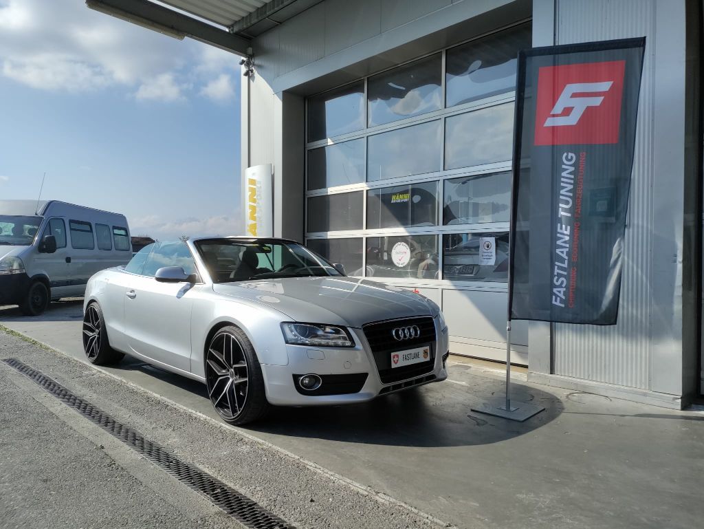 Ein perlweisses Audi A5 2.0 TFSI Cabriolet mit geschlossenem Dach steht vor der Garage neben der Fastlane Tuning Schweiz Flagge