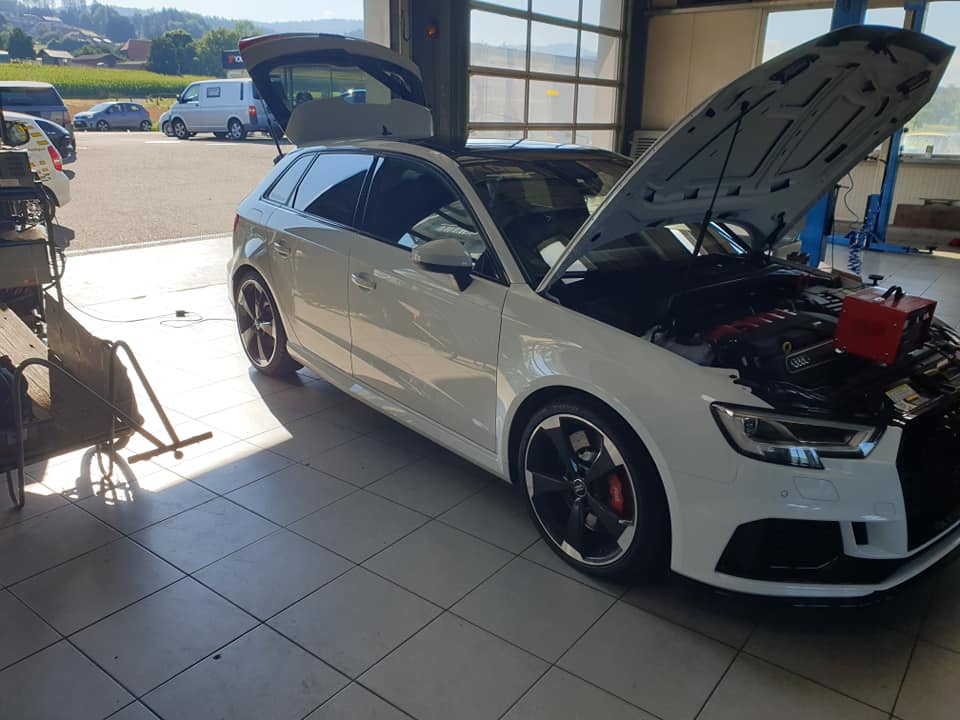 Ein weißer Audi RS3 steht mit geöffneter Motorhaube in der Garage, an der ein Batterieerhaltungsgerät angebracht ist