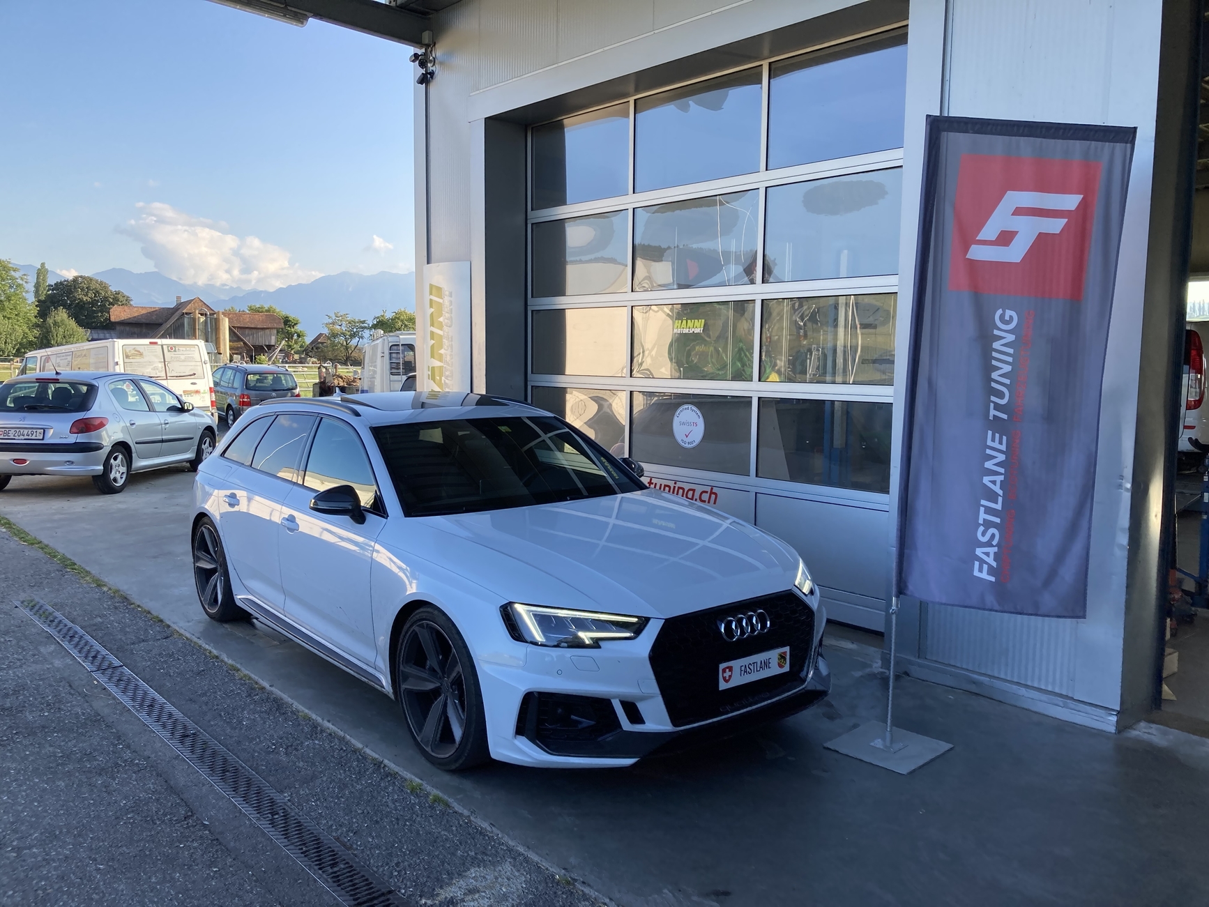 Ein weißer Audi RS4 V6 Kombi vor der Garage neben der Fastlane Tuning Schweiz Flagge