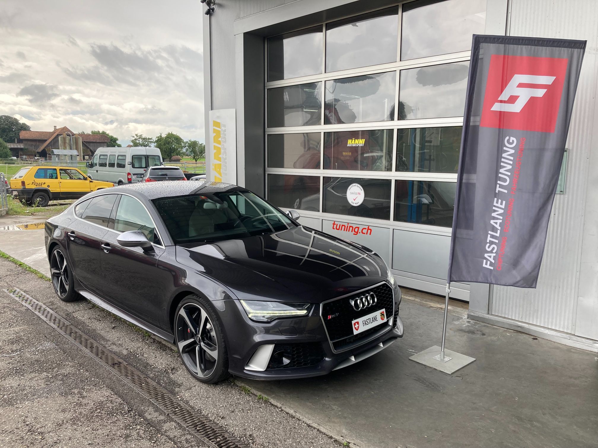 Ein schwarzer Audi RS7 steht vor der Garage neben der Fastlane Tuning Schweiz Flagge