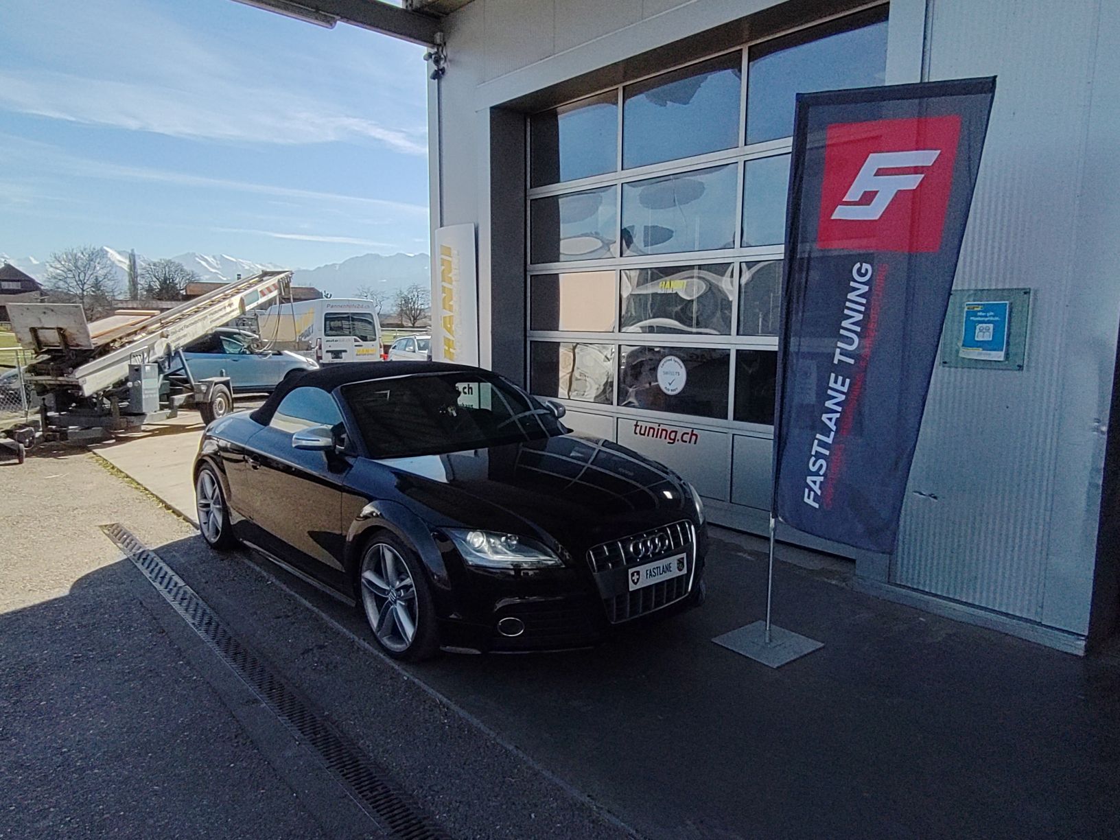 Ein schwarzes Audi TTS Mark 1 Cabriolet, geparkt vor der Garage neben der Fastlane Tuning Schweiz Flagge. Das Dach ist oben.