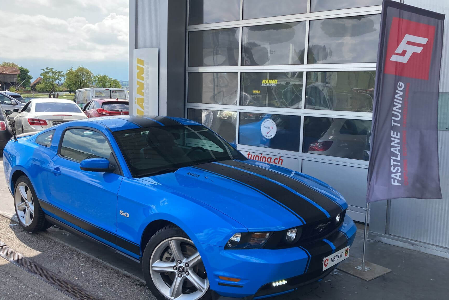 Ein himmelblauer Mustang GT steht neben der Fastlane Tuning Schweiz Flagge