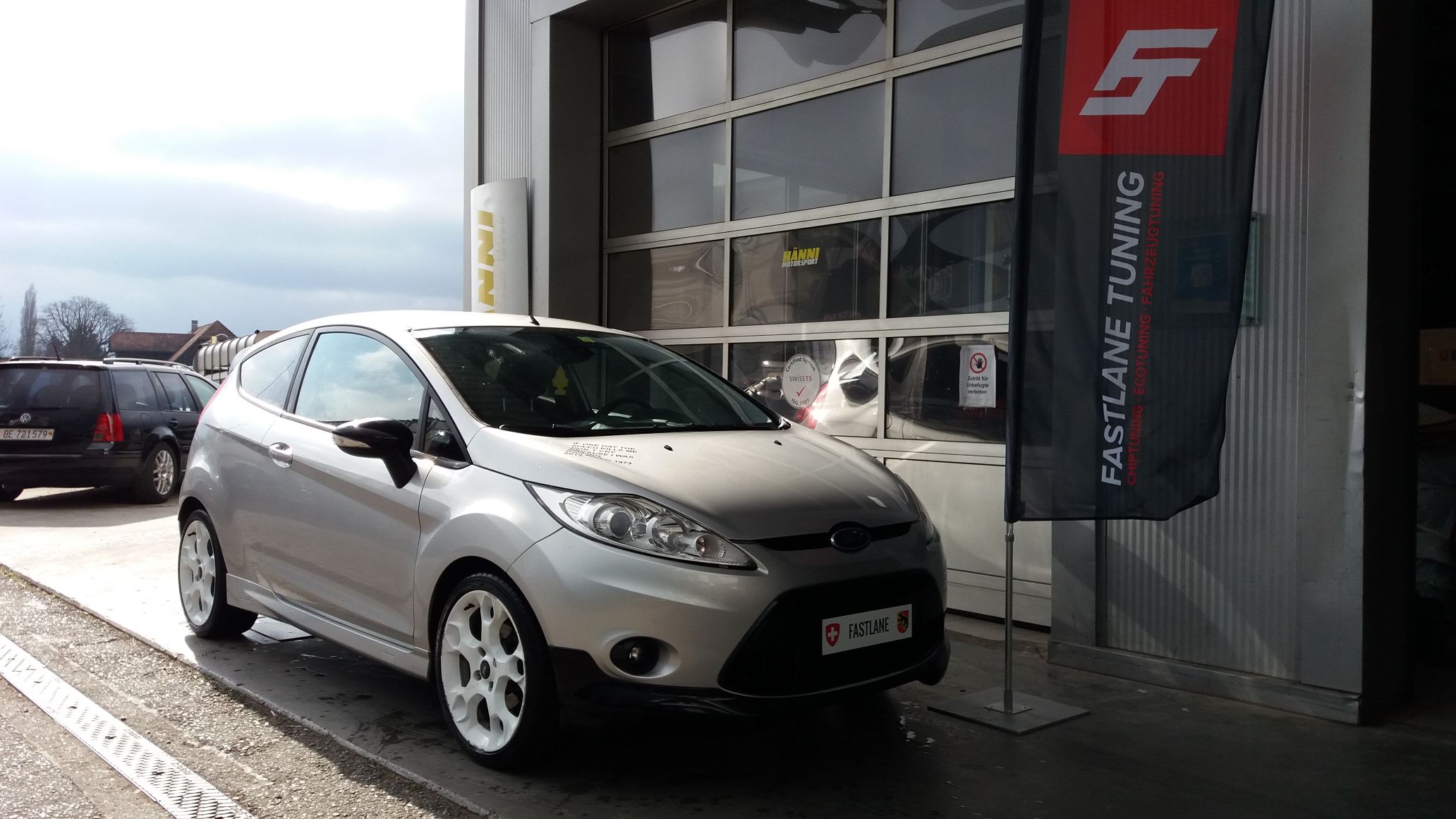 Ein kleiner silberner Ford Fiesta Mark 7 mit weissen Felgen steht vor der Garage neben der Fastlane Tuning Schweiz Flagge