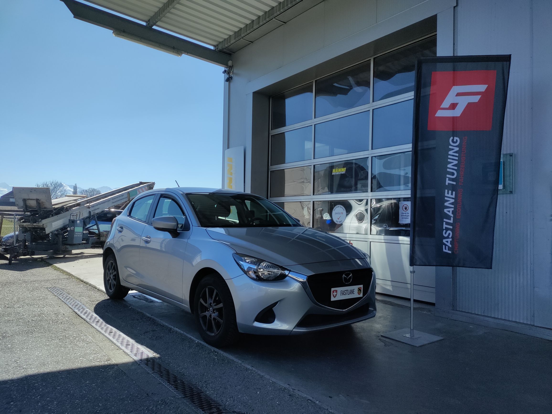 Ein silberner Mazda 2 skyaktiv 1.5 Liter steht vor der Garage neben der Fastlane Tuning Schweiz Flagge