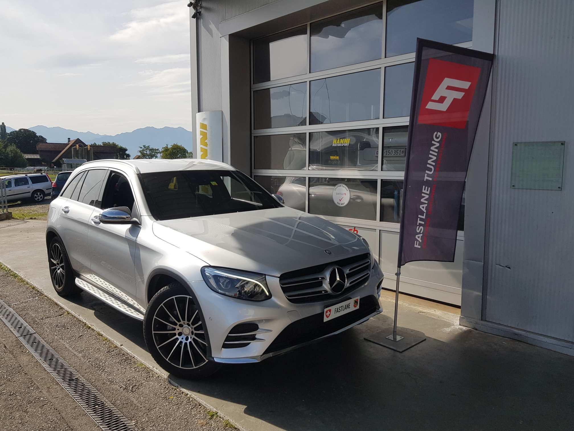 Ein silberner Mercedes Benz GLC 250d parkt vor der Garage neben der Fastlane Tuning Schweiz Flagge