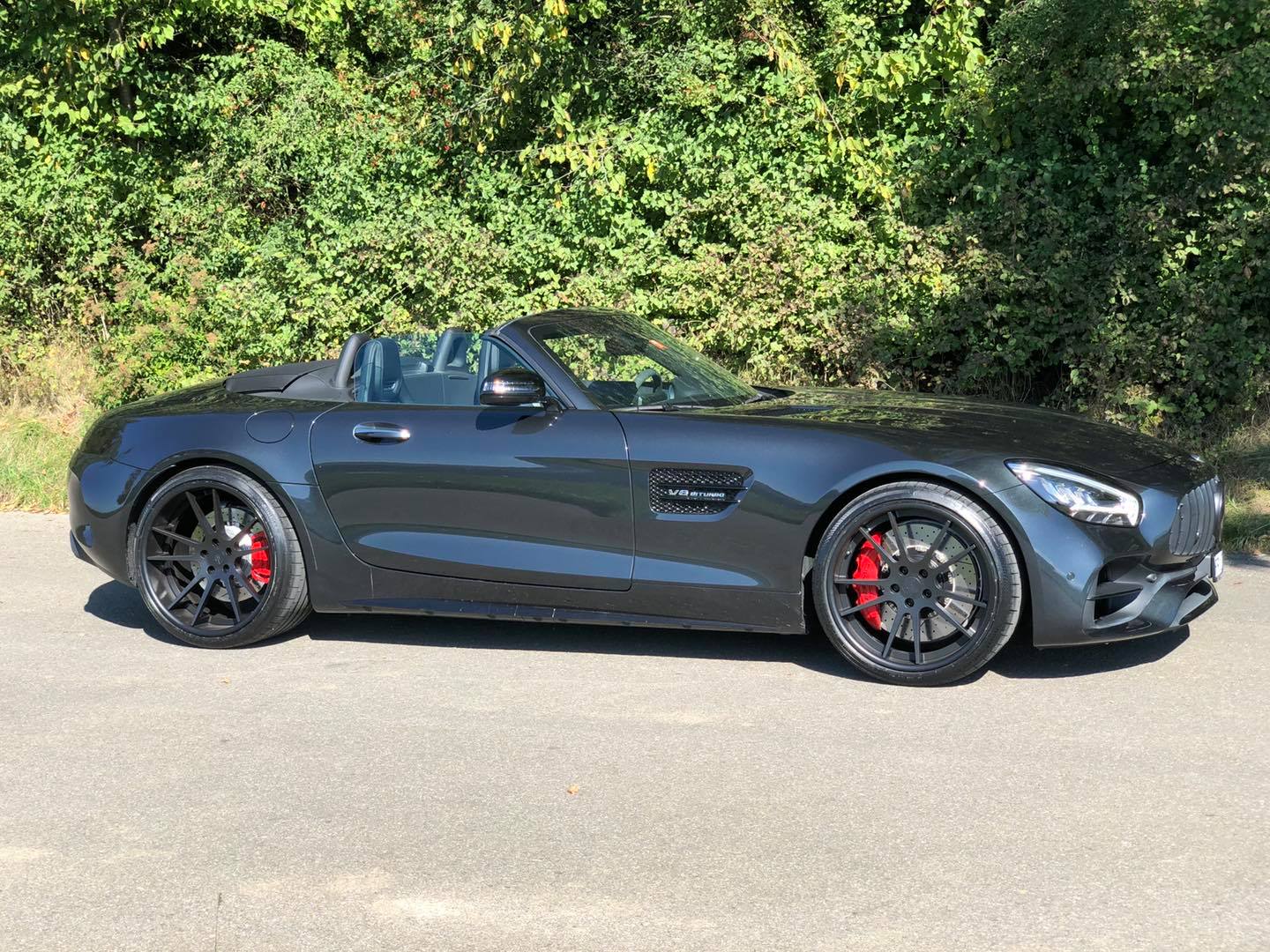 Ein kohlegrauer Mercedes AMG GTC Roadster mit schwarzen Rädern steht auf einem hellbraunen Feldweg in der Nähe von Bäumen.