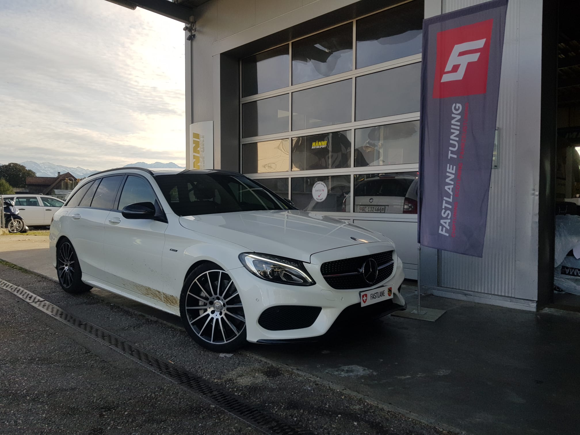 Ein weisser Mercedes C450 AMG mit silbernen Felgen steht neben der fastlane tuning schweiz Flagge