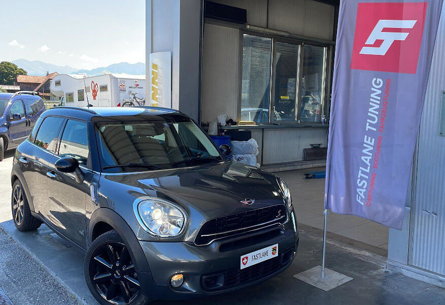 Ein anthrazitfarbener Mini Countryman Cooper S steht neben der Fastlane Tuning Schweiz Flagge.