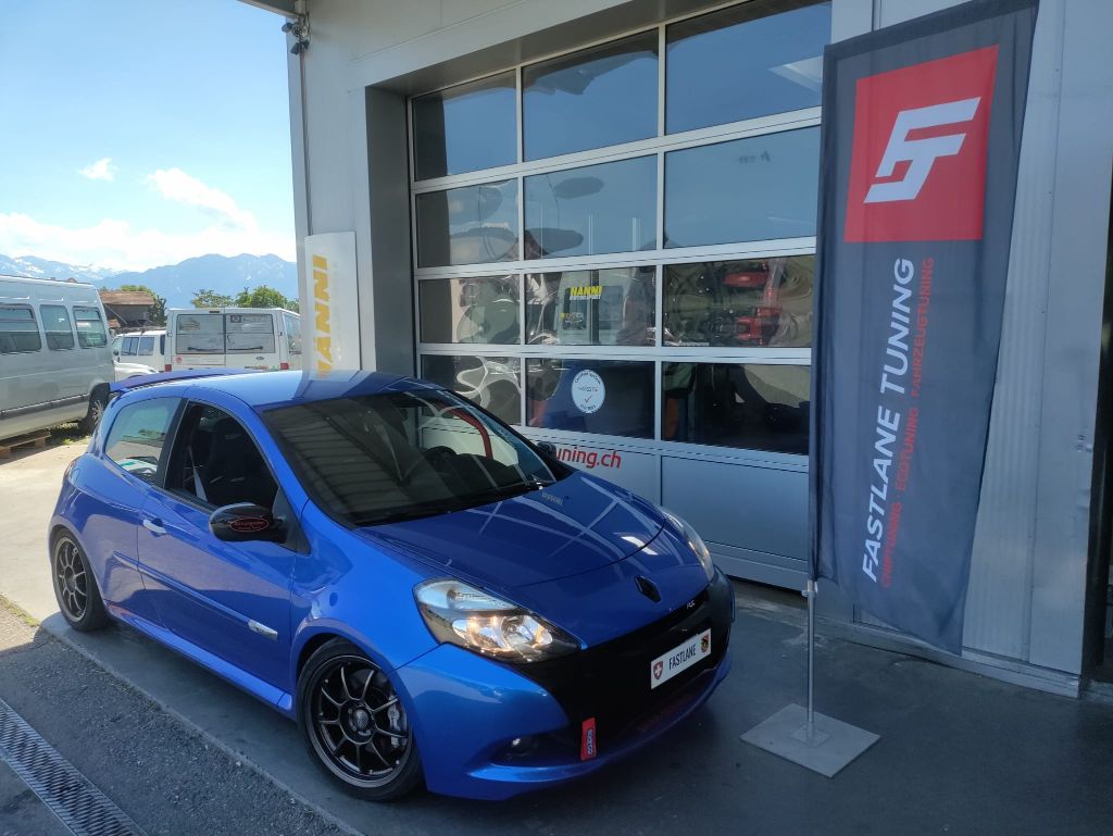 Ein dunkelblauer Renault Clio III RS Trophy Edition, der vor der Garage neben der Fastlane Tuning Schweiz Flagge geparkt ist