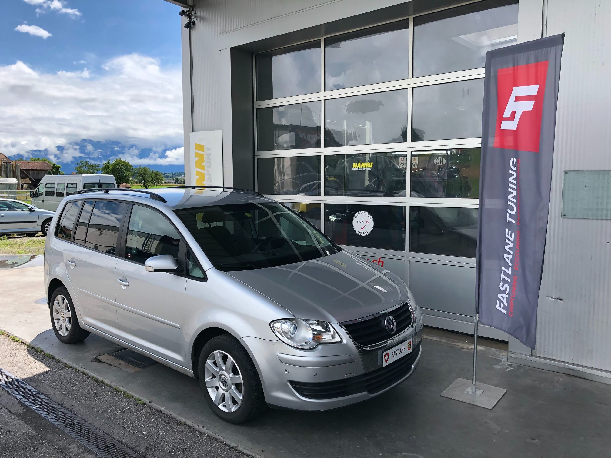 Ein grauer VW Touran steht neben der Fastlane Tuning Schweiz Flagge