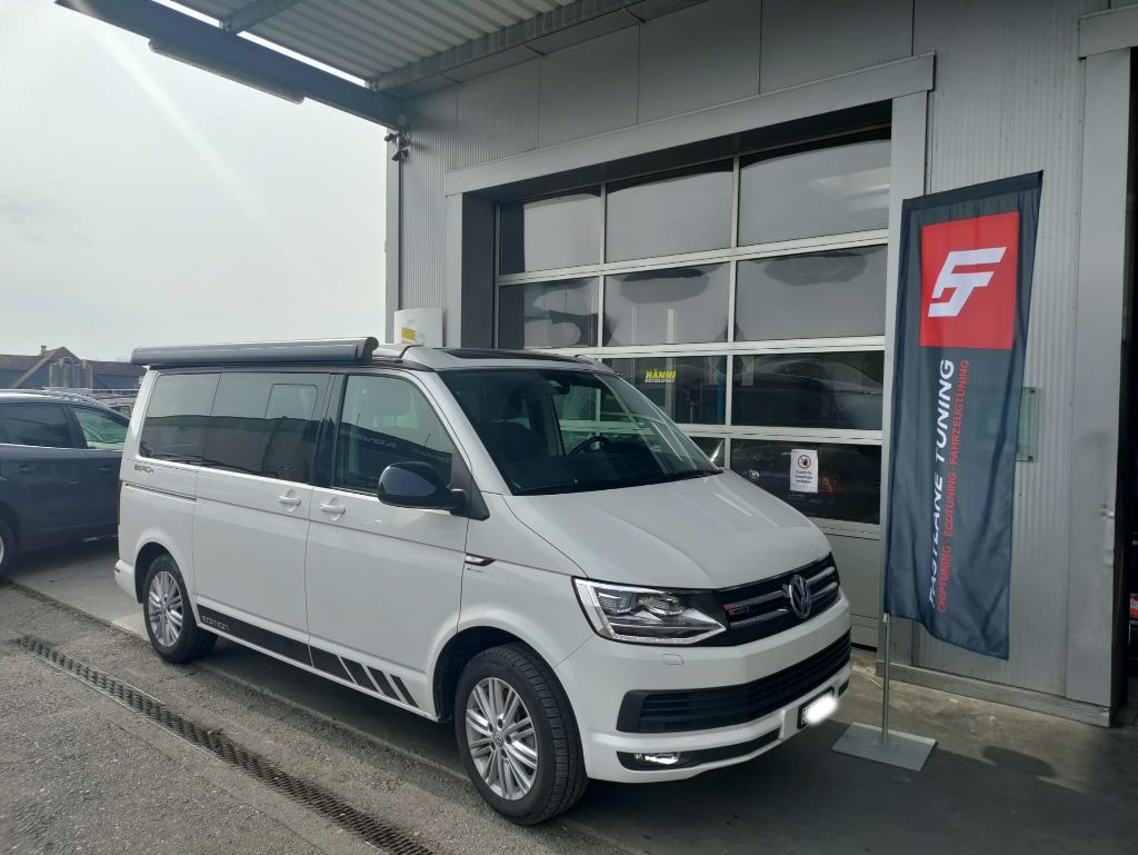 Ein weisser Volkswagen T6 Multivan 2.0 TDI parkt vor der Garage neben der Fastlane Tuning Schweiz Flagge