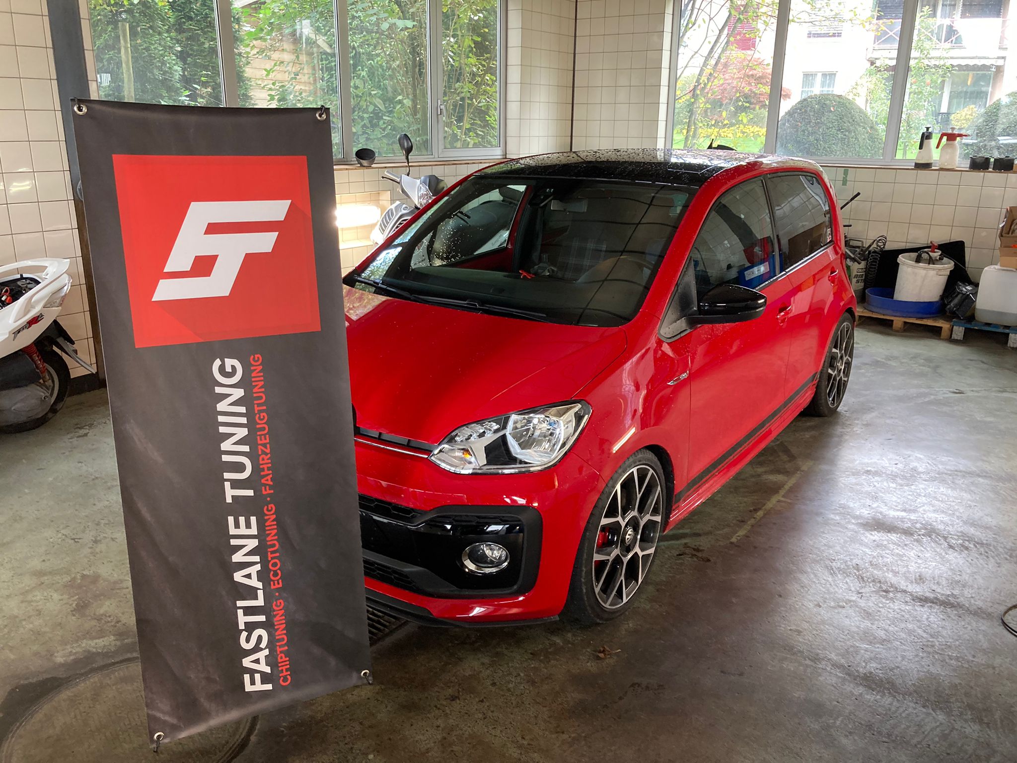 Ein roter Volkswagen Up steht neben dem Banner von Fastlane Tuning Schweiz in einer Waschanlage.