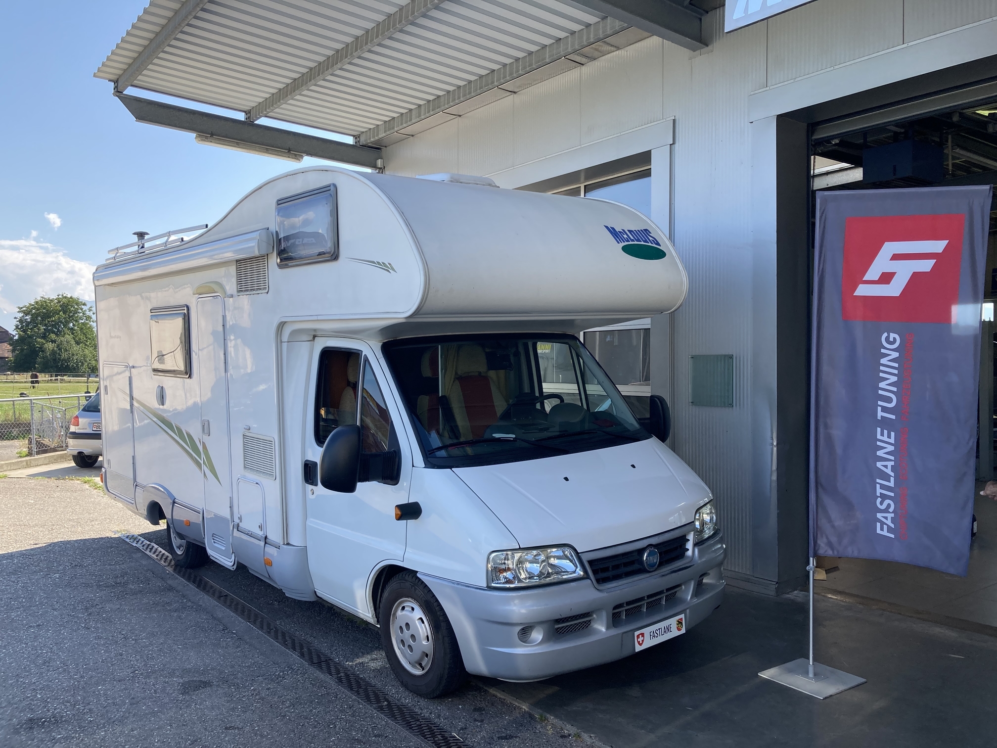 Ein weisser Fiat Ducato II 2.3 Multijet steht vor der Garage neben der Fastlane Tuning Schweiz Flagge