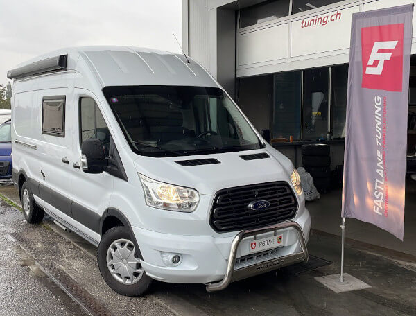 Ein weisses Wohnmobil auf Ford Transit Basis steht neben der Fastlane Tuning Schweiz Flagge.