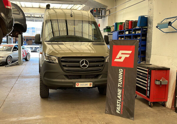 Ein olivgrünes Mercedes Sprinter 907 AWD Wohnmobil steht neben dem Banner der Fastlane Tuning Schweiz GmbH.