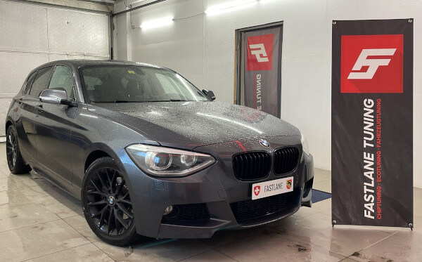 Ein grauer BMW 118i steht neben dem Banner der Fastlane Tuning Schweiz GmbH