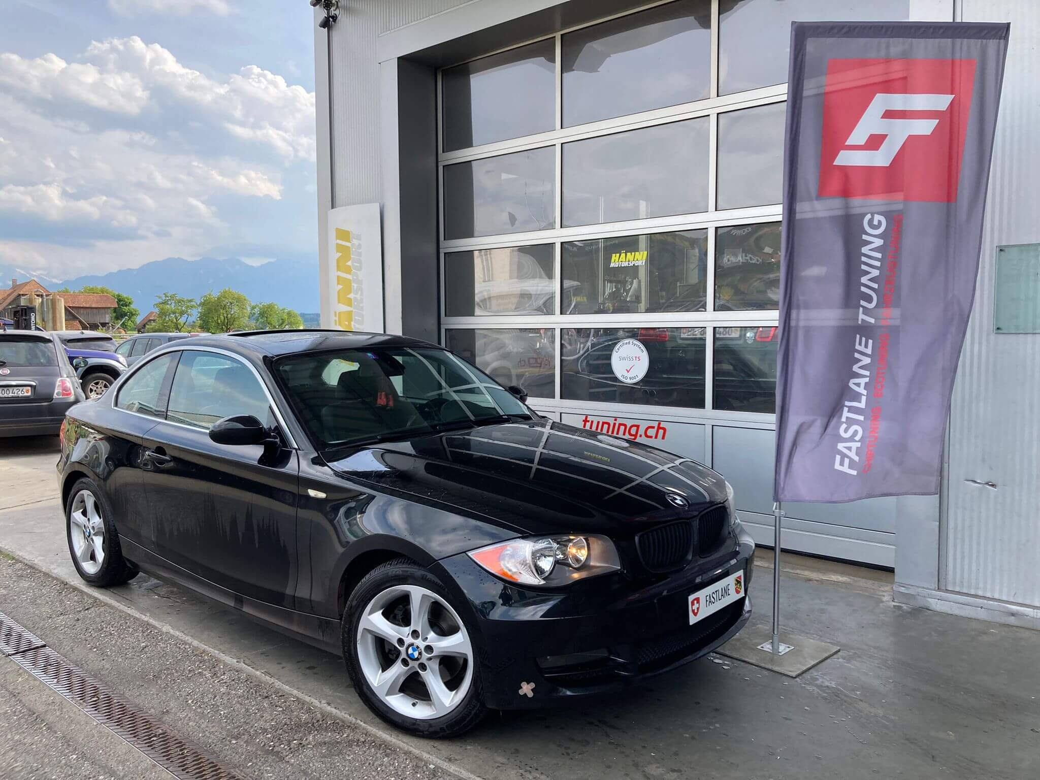 Ein schwarzes BMW 128i Coupé steht neben der Fastlane Tuning Schweiz Flagge. Es handelt sich um eine spezielle US-Version, die für die kalifornischen Abgasvorschriften SULEV II ausgelegt ist.