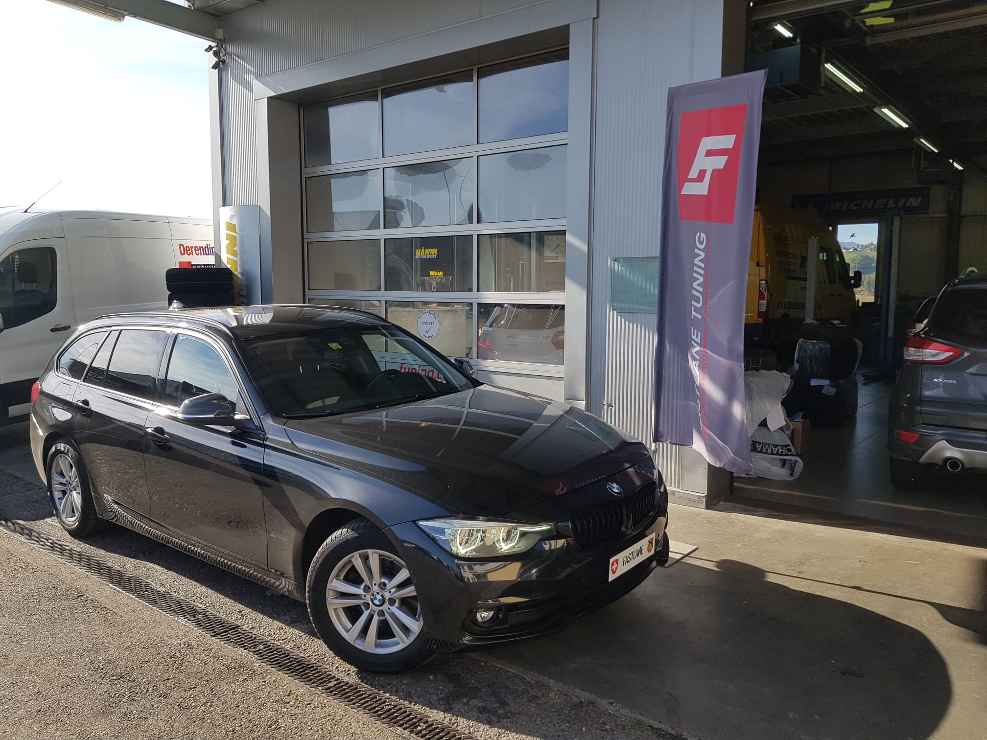 Ein schwarzer BMW 320d Kombi steht neben der fastlane tuning schweiz Flagge