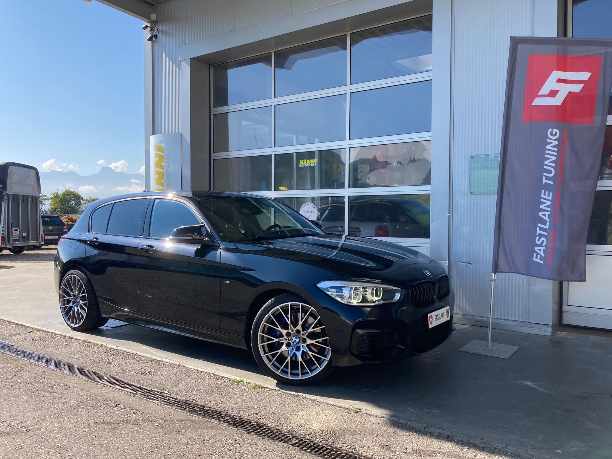 Ein dunkelschwarzer BMW M140i mit Stage 2 Performance Tuning und silbernen Felgen parkt neben der Fastlane Tuning Schweiz Flagge.